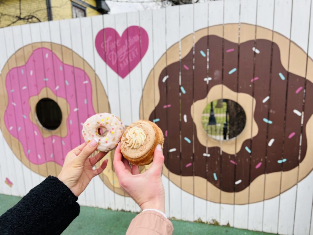 Erinn and Olivia hold donuts in front of a donut mural at Five Daughters Bakery.
